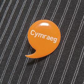 orange speech bubble shaped badge on a pinstriped piece of fabric. Text on the badge says "Cymraeg"