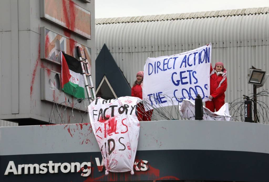 Photograph of two people in red boilersuits. They are standing on top of a factory, there is red paint everywhere, a Palestinian flag, and homemade banners reading "This factory kills kids" and "direct action gets the goods"