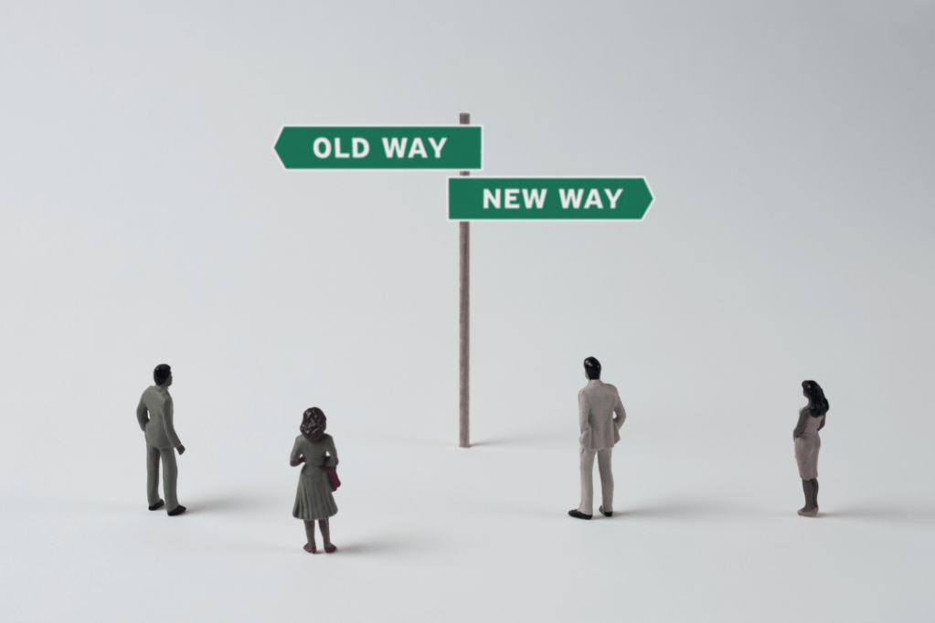 miniature model people looking at a crossroads sign, one reads Old Way, one reads new way